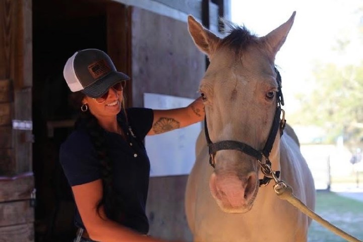 Person in a cap petting a light-colored horse near a stable entrance.