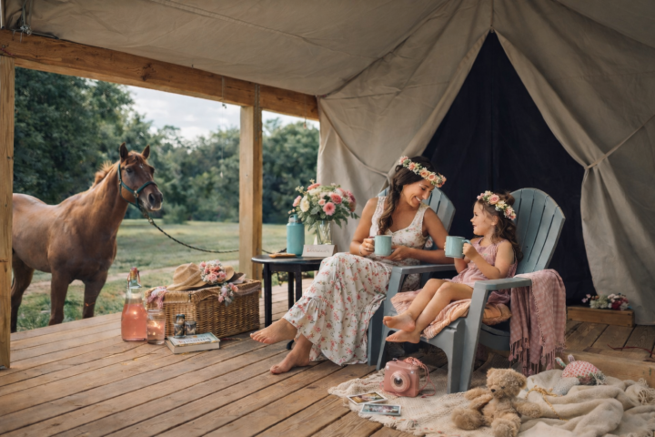 Woman and girl wearing flower crowns sit in chairs on porch near horse, sipping from mugs.